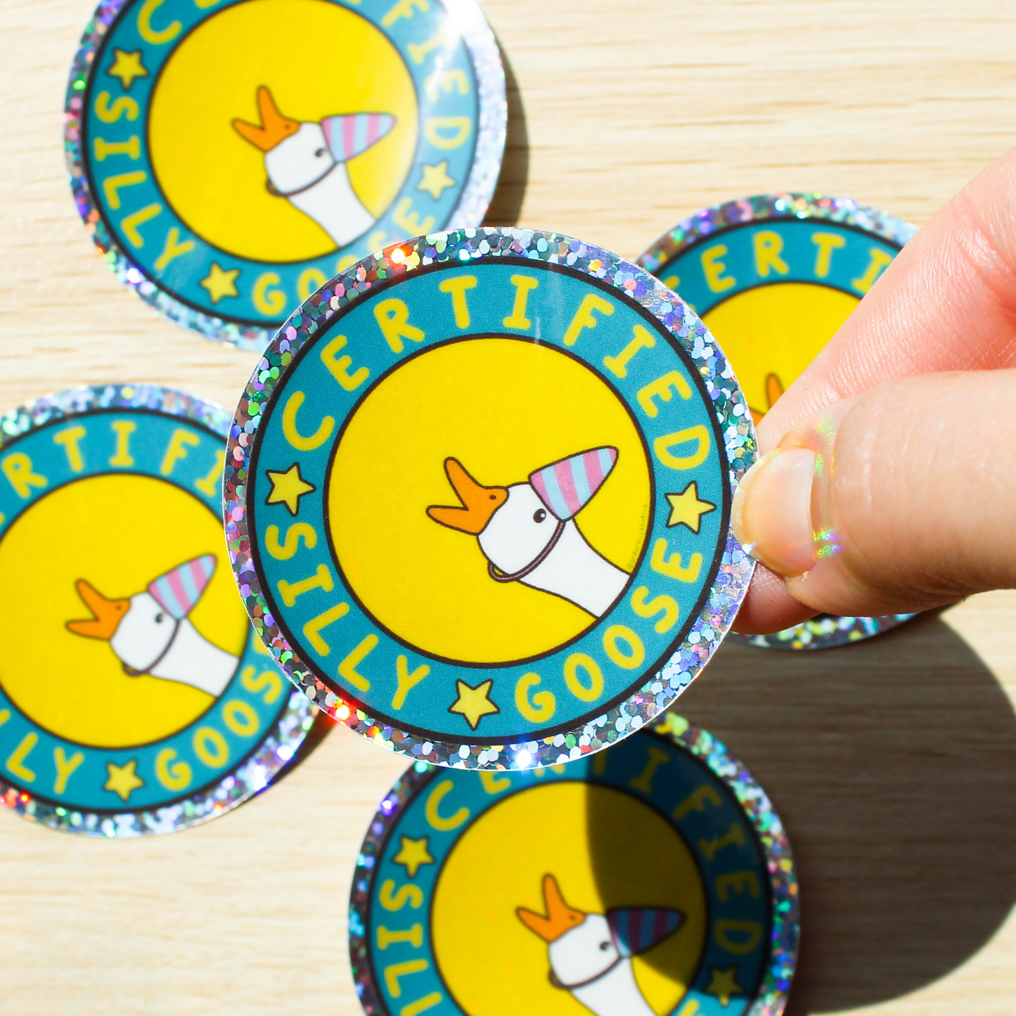 Hand holding a holographic glitter sticker with Certified Silly Goose Sticker written in a circle with stars in between the words. A goose with a blue and pink party hat is popping out on a yellow background. The background has more silly goose stickers on a light wood background
