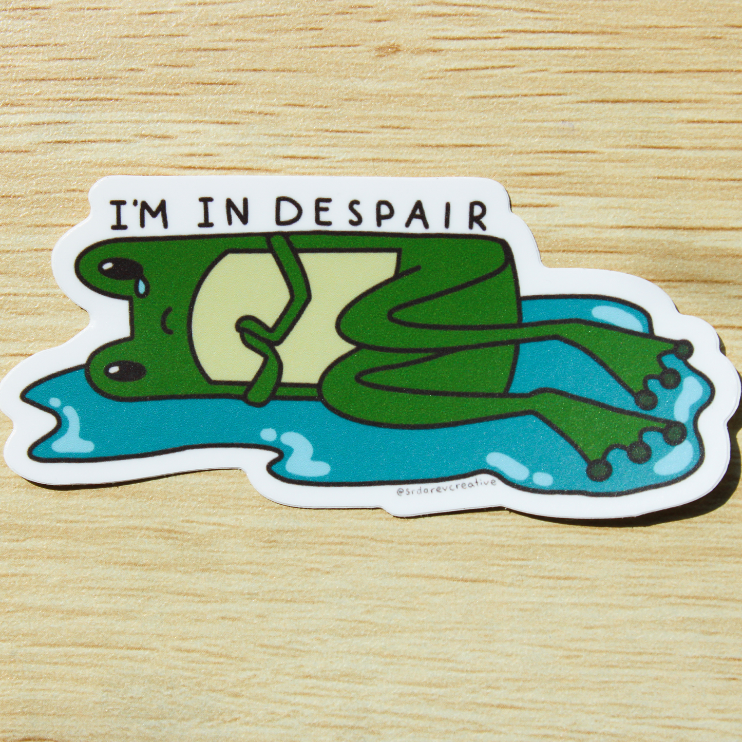 Hand holding a sticker of a cartoon frog lying down in a puddle with a tear falling down one eye and holding its hands. On top is written "I'M IN DESPAIR". Background is a wooden table.