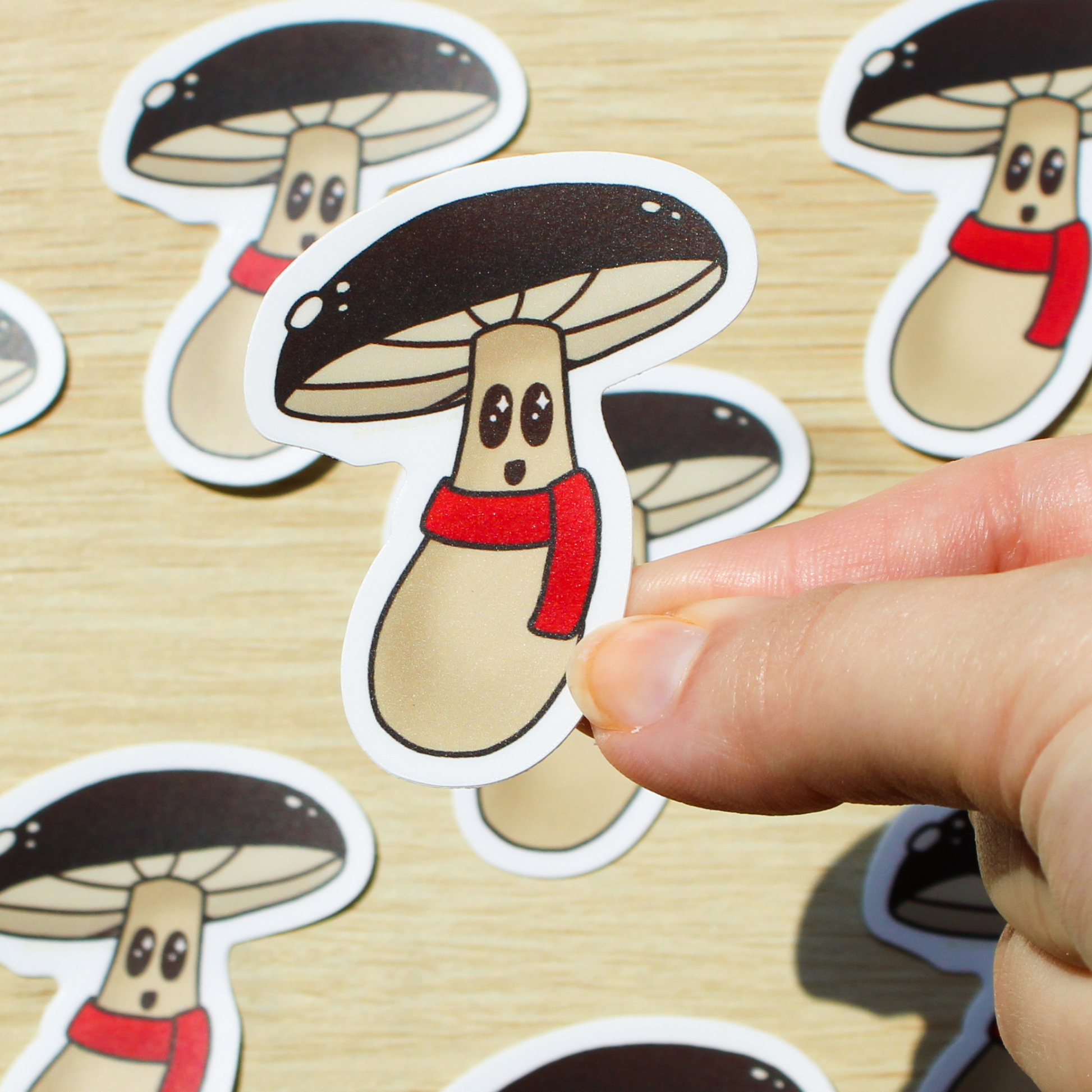 A wooden background with multiple mushroom stickers across it. In the front is a hand holding a mushroom sticker with a brown cap, smiling face, and a red scarf.