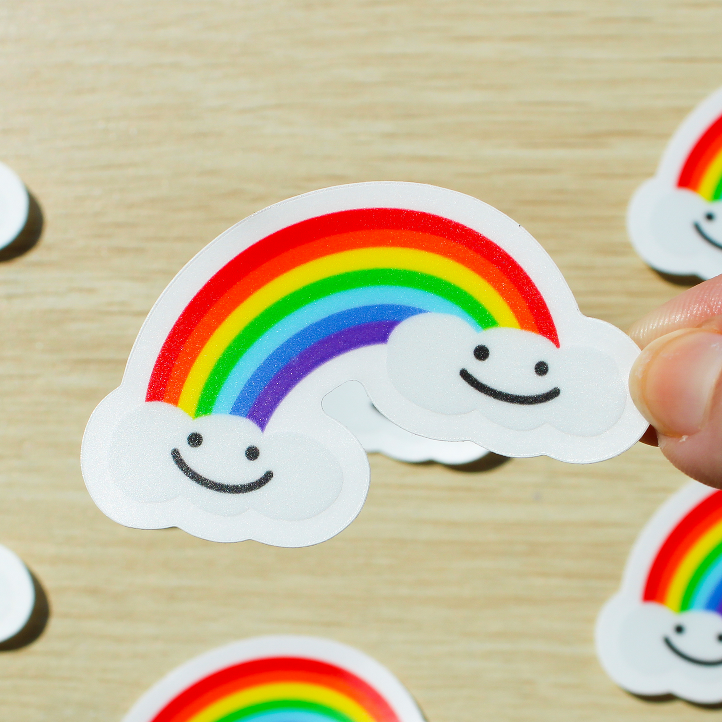 A wooden background with rainbow stickers on the ground. In the front is a hand holding a sticker that has a rainbow with two clouds on either end smiling.