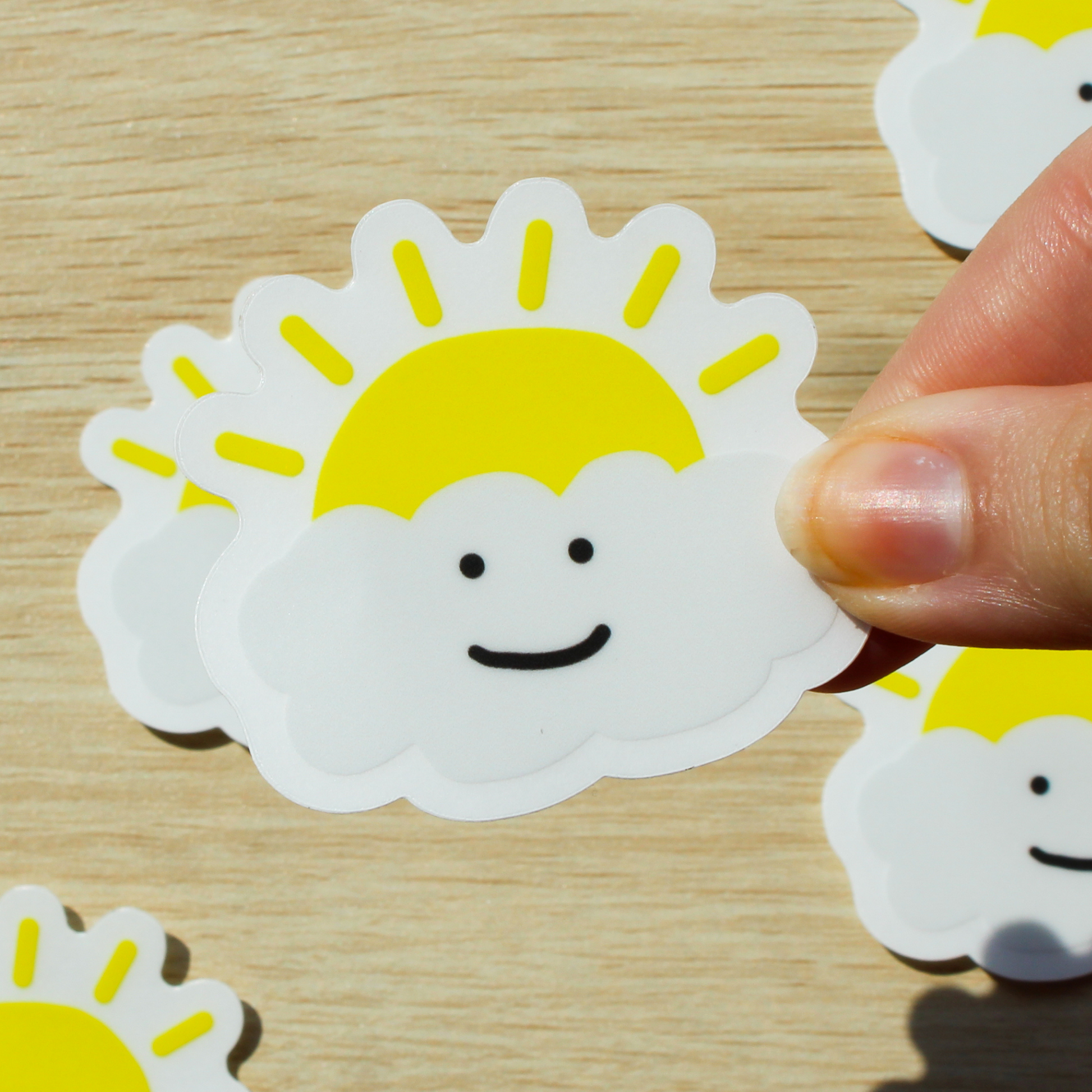 Background is wood with more happy cloud stickers. In front is a hand holding a happy cloud sticker with a sun popping out behind it.