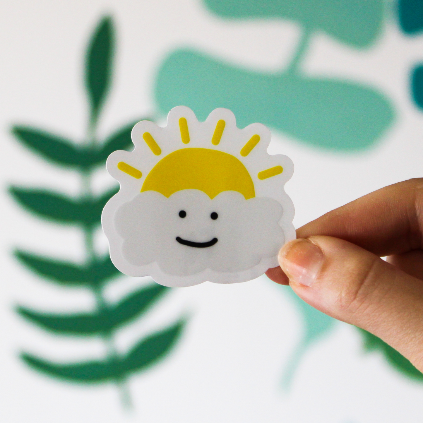 A blurred leaf pattern in the background. In front is a hand holding a happy cloud sticker with a sun popping out behind it.