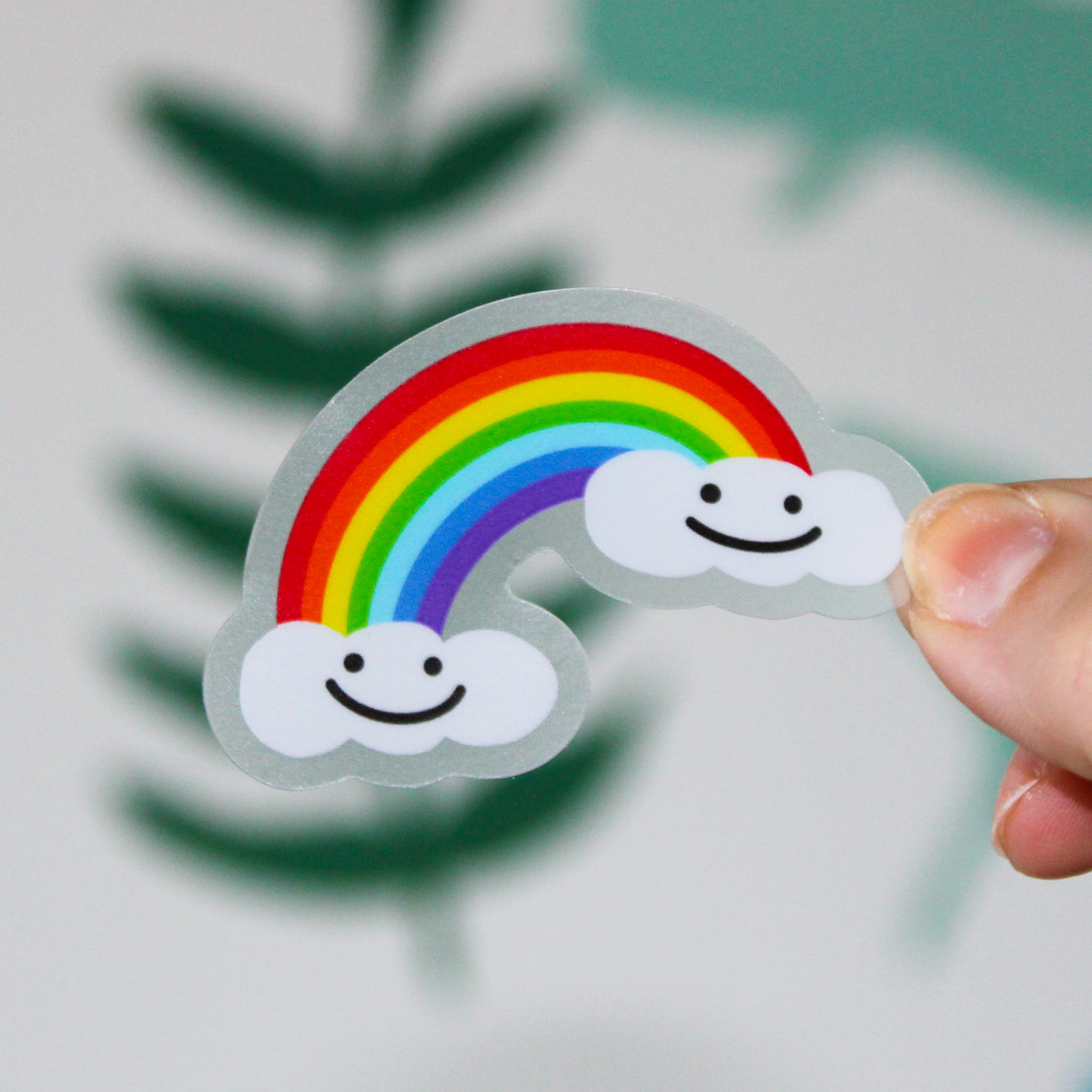 A blurred leaf background. In the front is a hand holding a sticker that has a rainbow with two clouds on either end smiling.