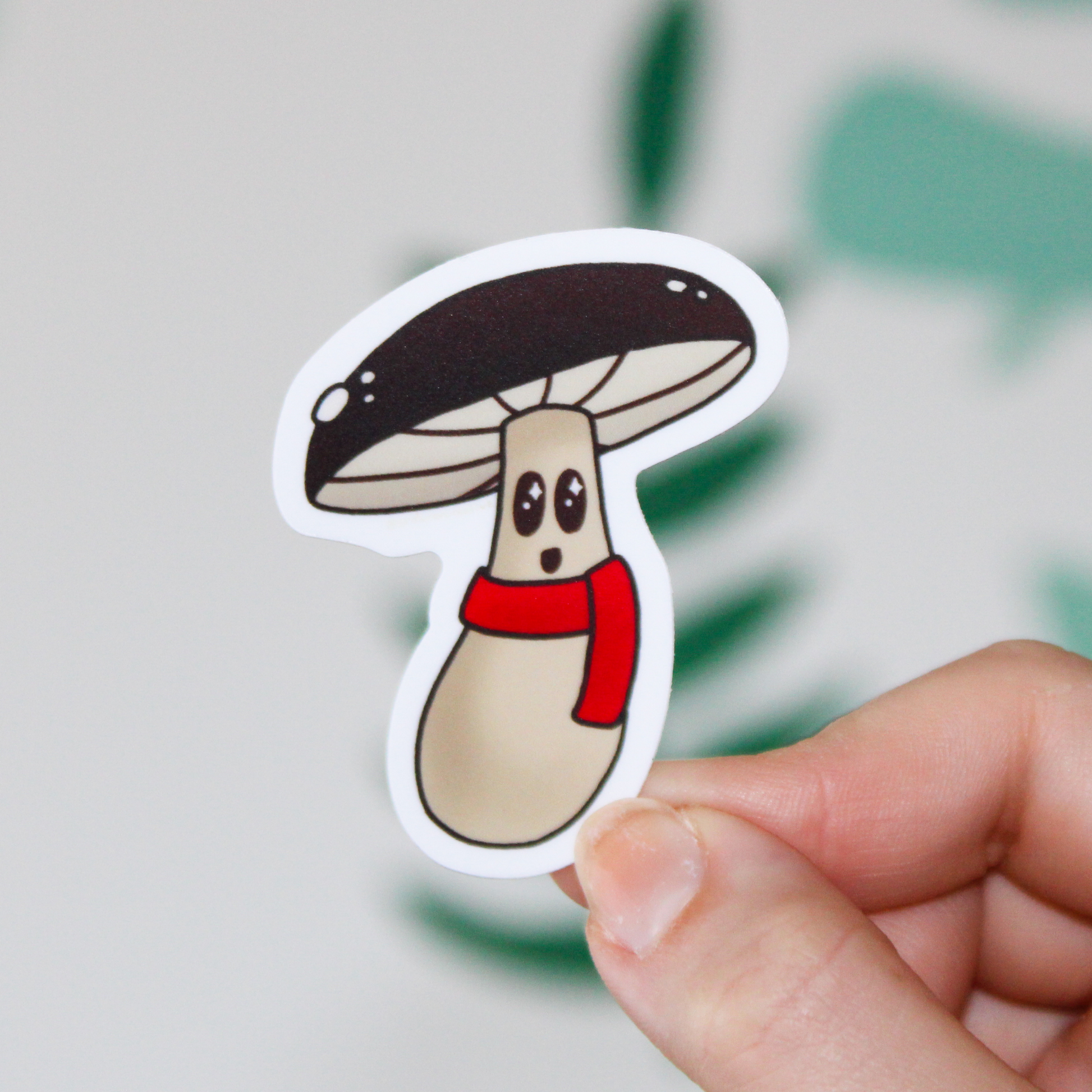 A blurred leaf background. In the front is a hand holding a mushroom sticker with a brown cap, smiling face, and a red scarf.