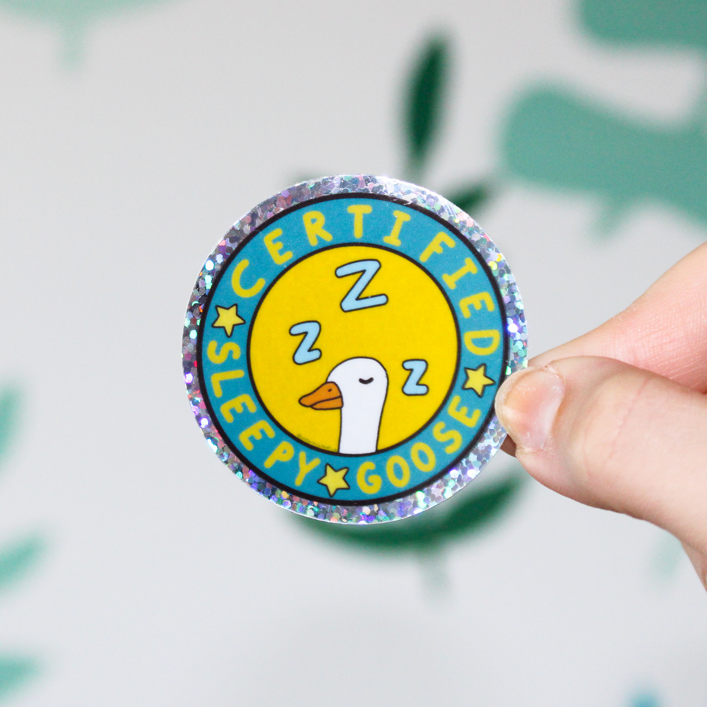 A hand holding a circle holographic sticker that has Certified Sleepy Goose written in a circle with stars and a white goose with Z's sleeping on a yellow background. Behind the sticker is a blurred leaf pattern.