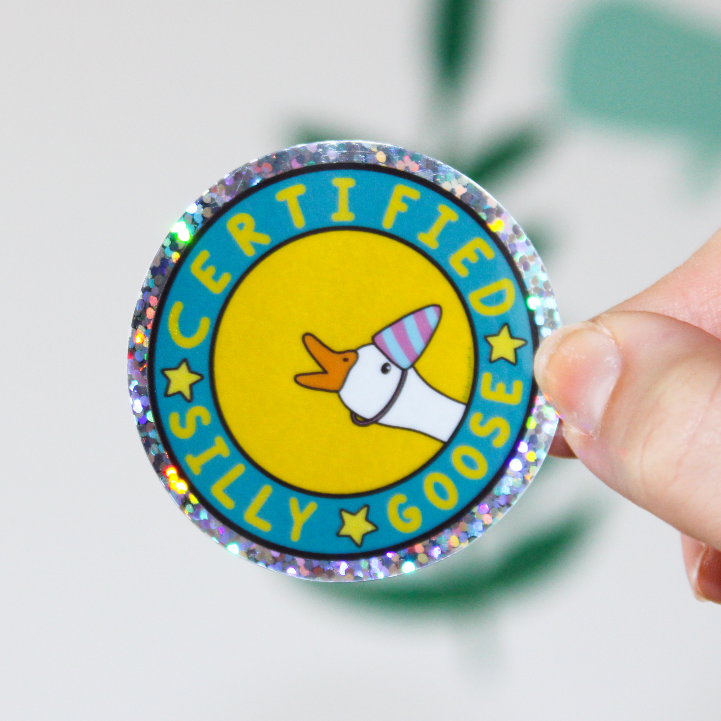 Hand holding a holographic glitter sticker with Certified Silly Goose Sticker written in a circle with stars in between the words. A goose with a blue and pink party hat is popping out on a yellow background