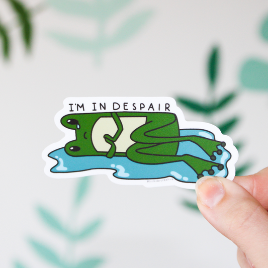 Hand holding a sticker of a cartoon frog lying down in a puddle with a tear falling down one eye and holding its hands. On top is written "I'M IN DESPAIR". Background is a blurred leaf pattern,