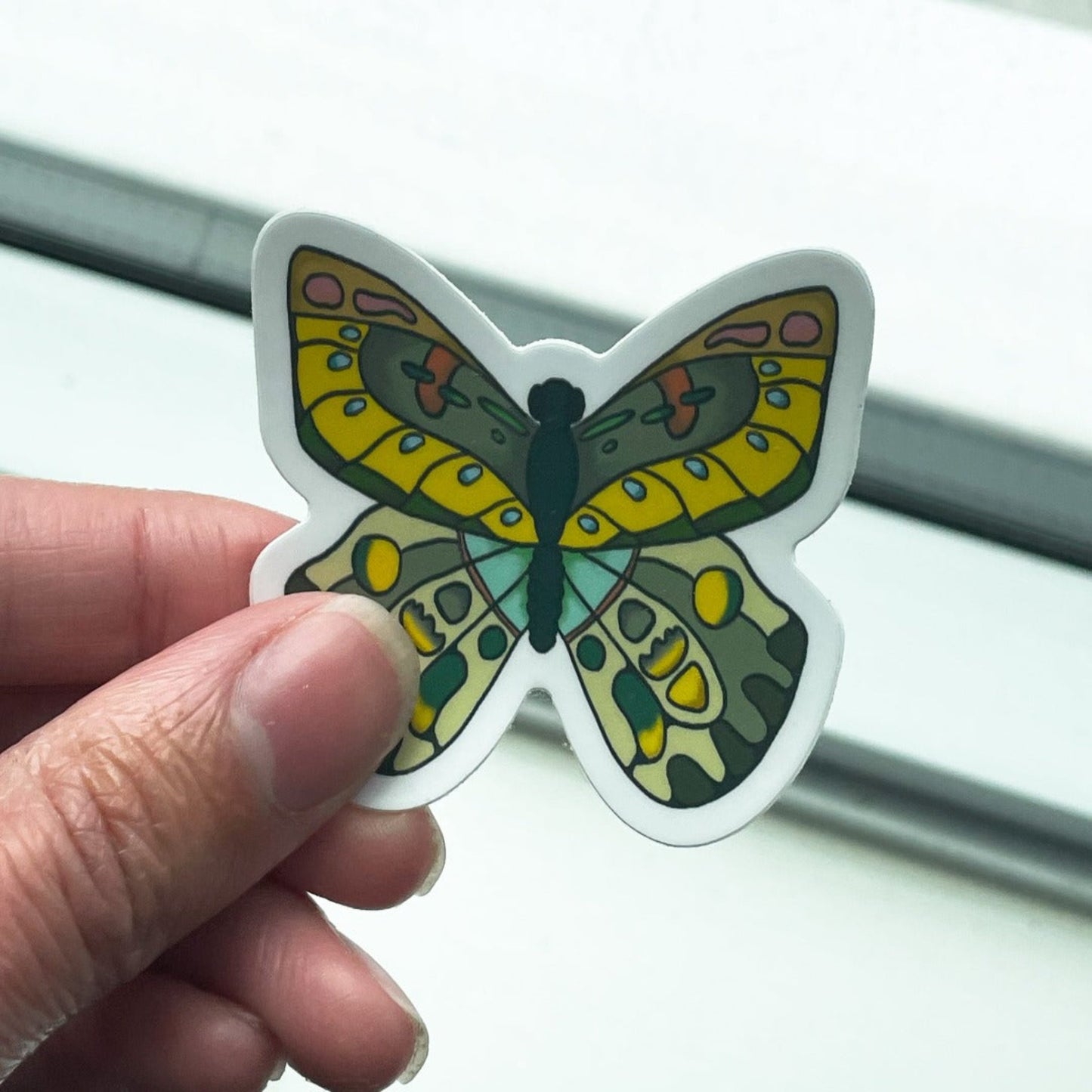 A butterfly sticker with blue, yellow, and green highlights is seen held in a hand on a white background