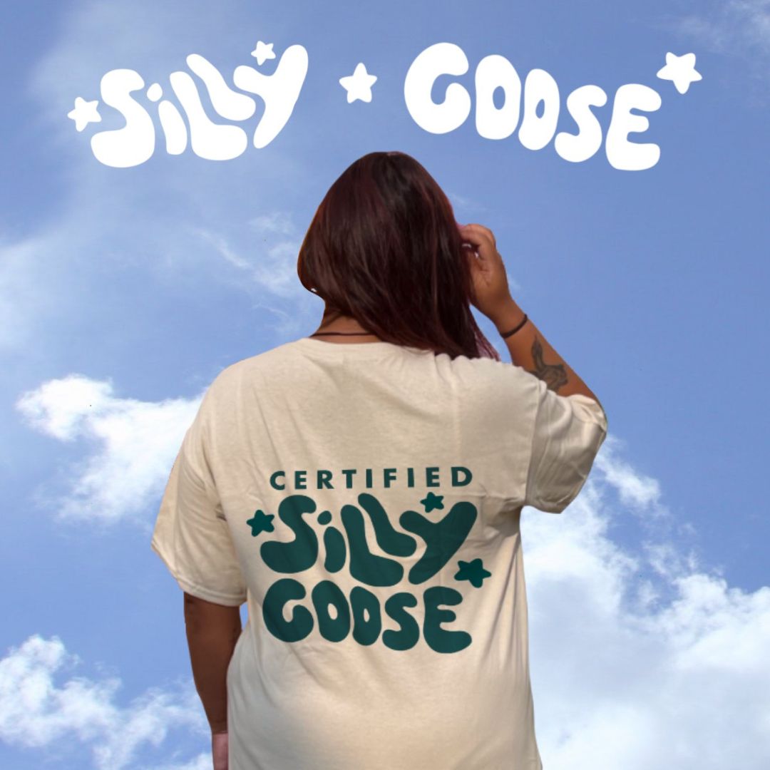 Blue sky background with clouds and "Silly Goose" written on top in white with stars. Below is the back of a woman wearing a beige shirt with "Certified Silly Goose" written and stars in dark blue.