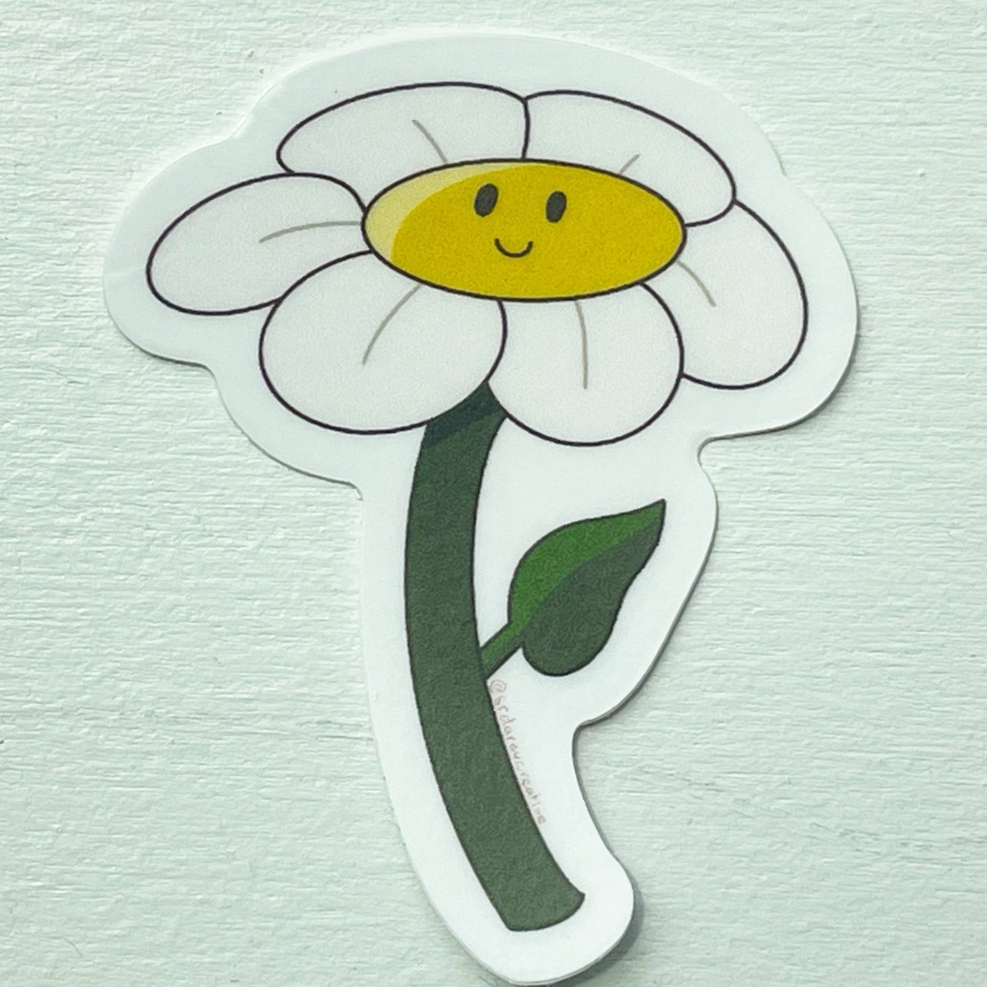 A white background with a flower sticker on top that has white petals, a dark green stem, and a yellow centre with a smiling face.
