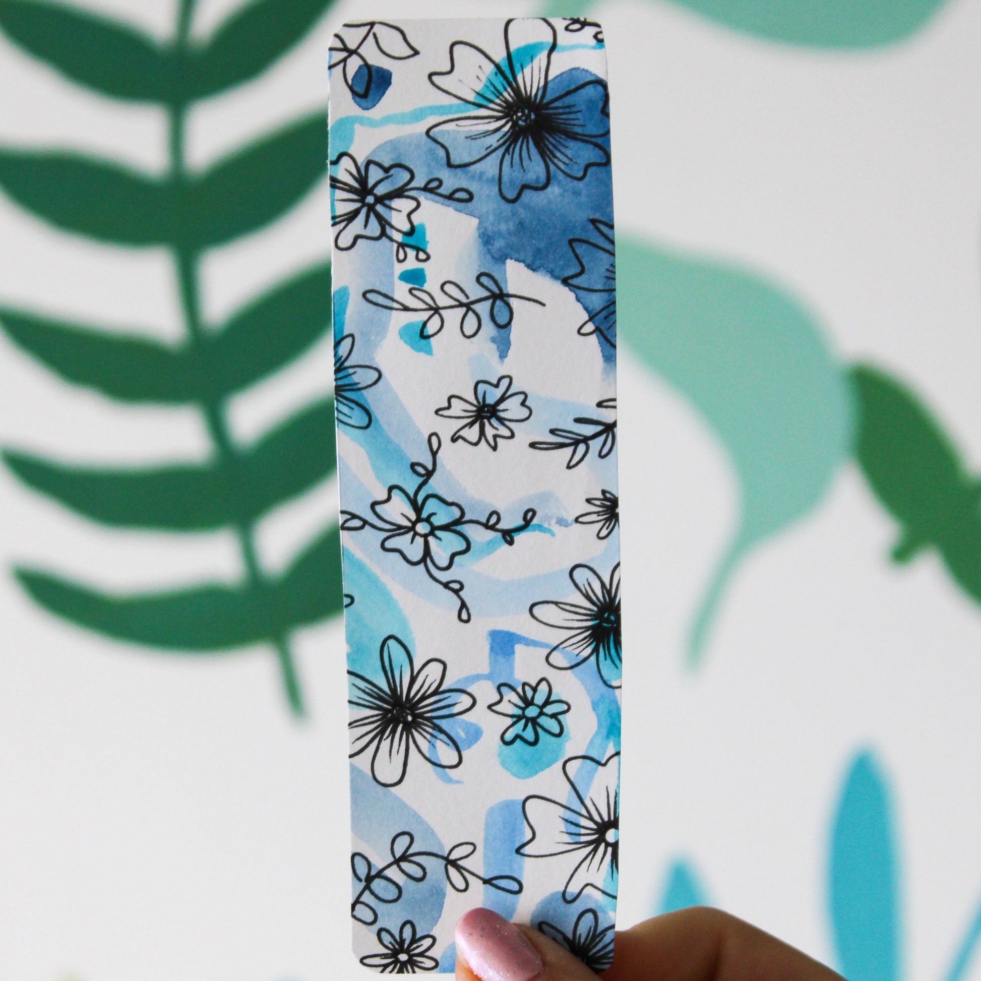 A blurred leaf pattern background. In the front is a hand holding a white and blue watercolour bookmark with black flowers and leaves drawn in ink.