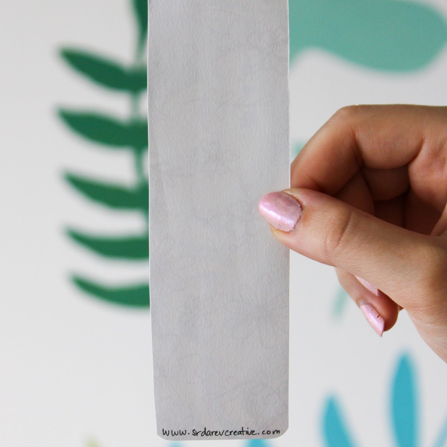 A blurred leaf background. In front is a hand holding a white bookmark and on the bottom is "www.srdarevcreative.com".