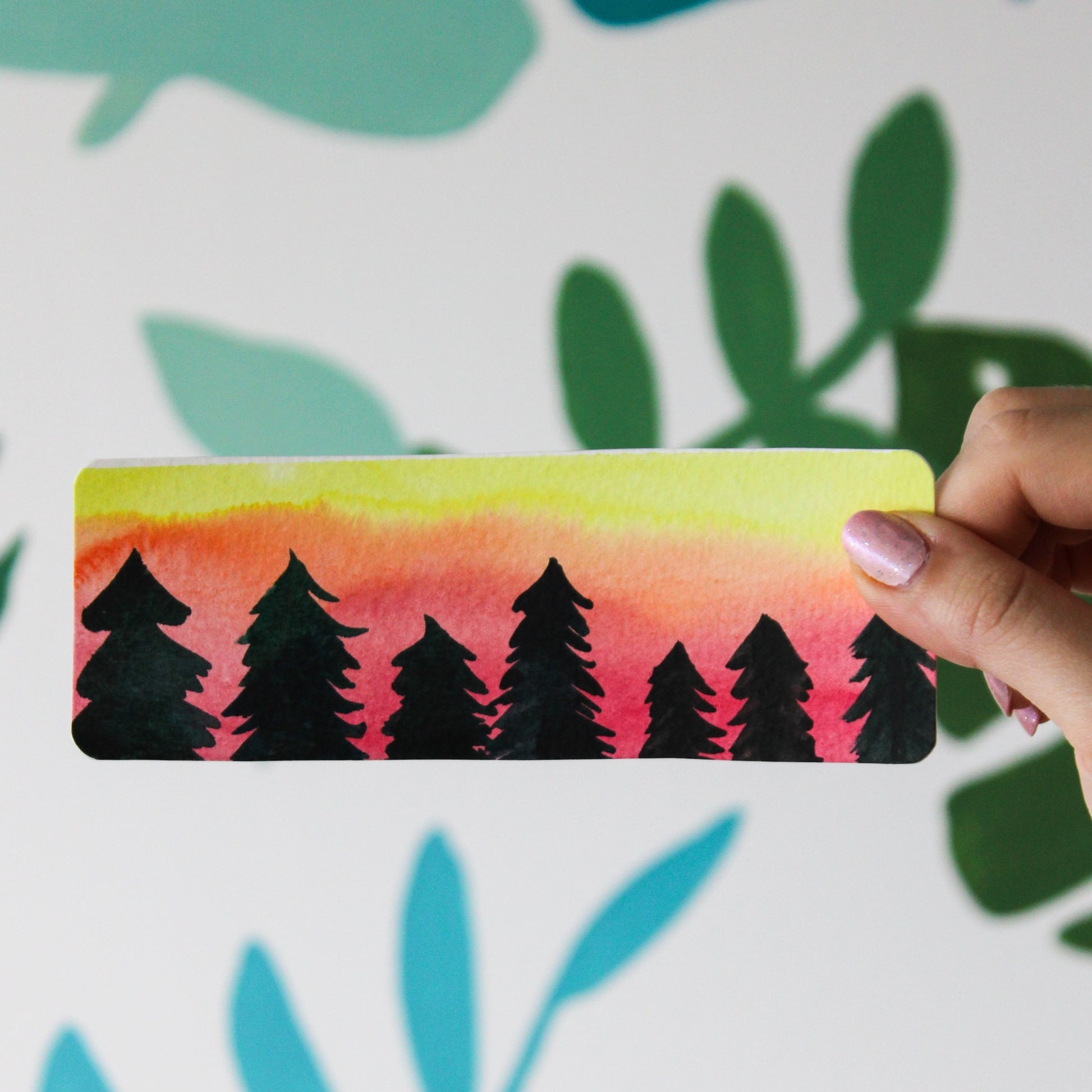 A blurred leaf background. In the front is a hand holding a hand painted bookmark with a watercolour sunset and black trees in the front.