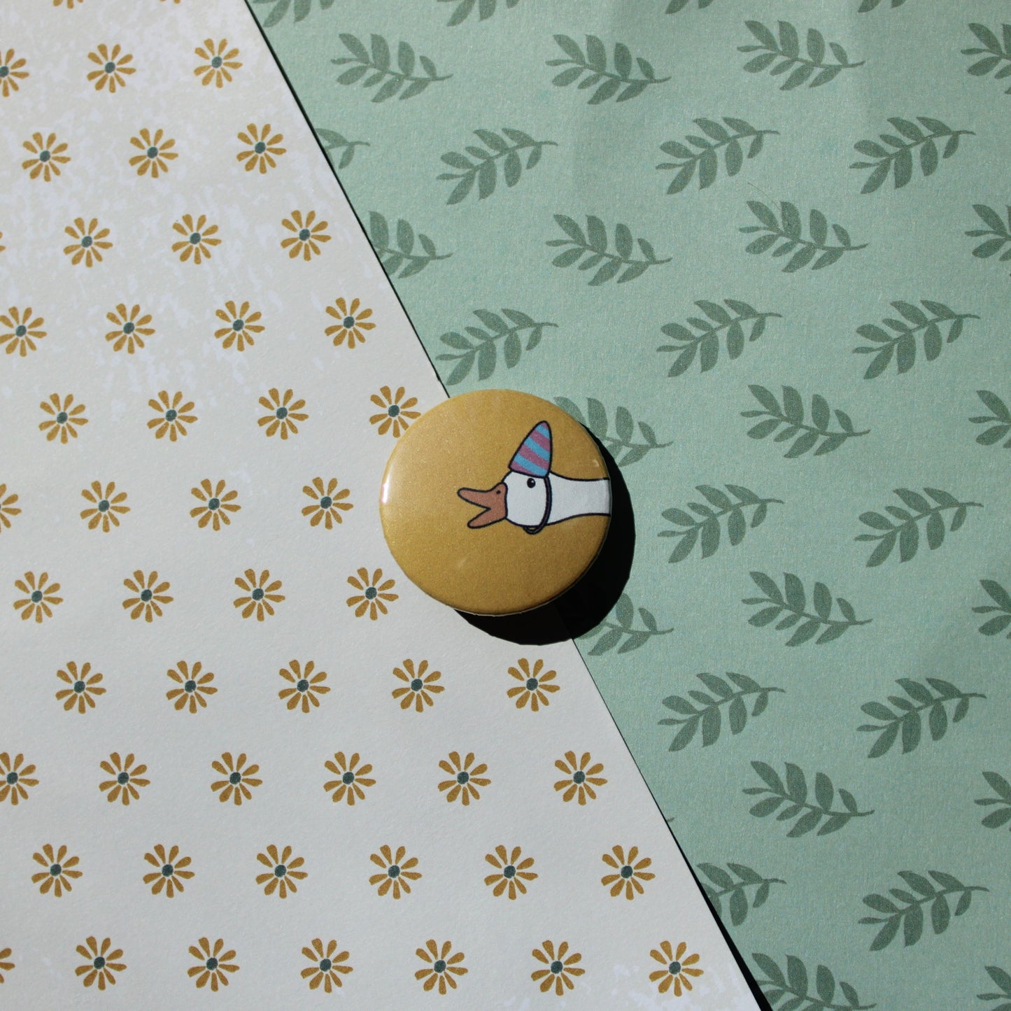 A floral and leaf background with a yellow circle button with a white goose head honking with a purple and blue striped party hat.
