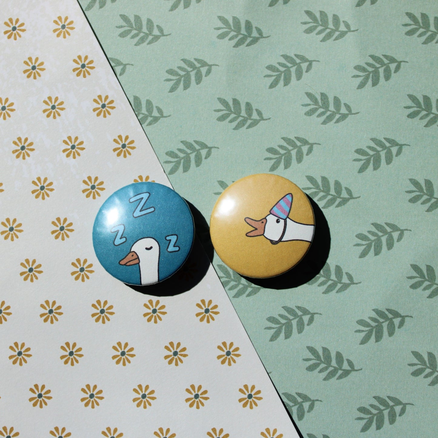 A floral and leaf background with one blue circle button with a white goose head sleeping and three Z's in light blue around it's head. Next to it is a yellow circle button with a white goose head honking with a purple and blue striped party hat.