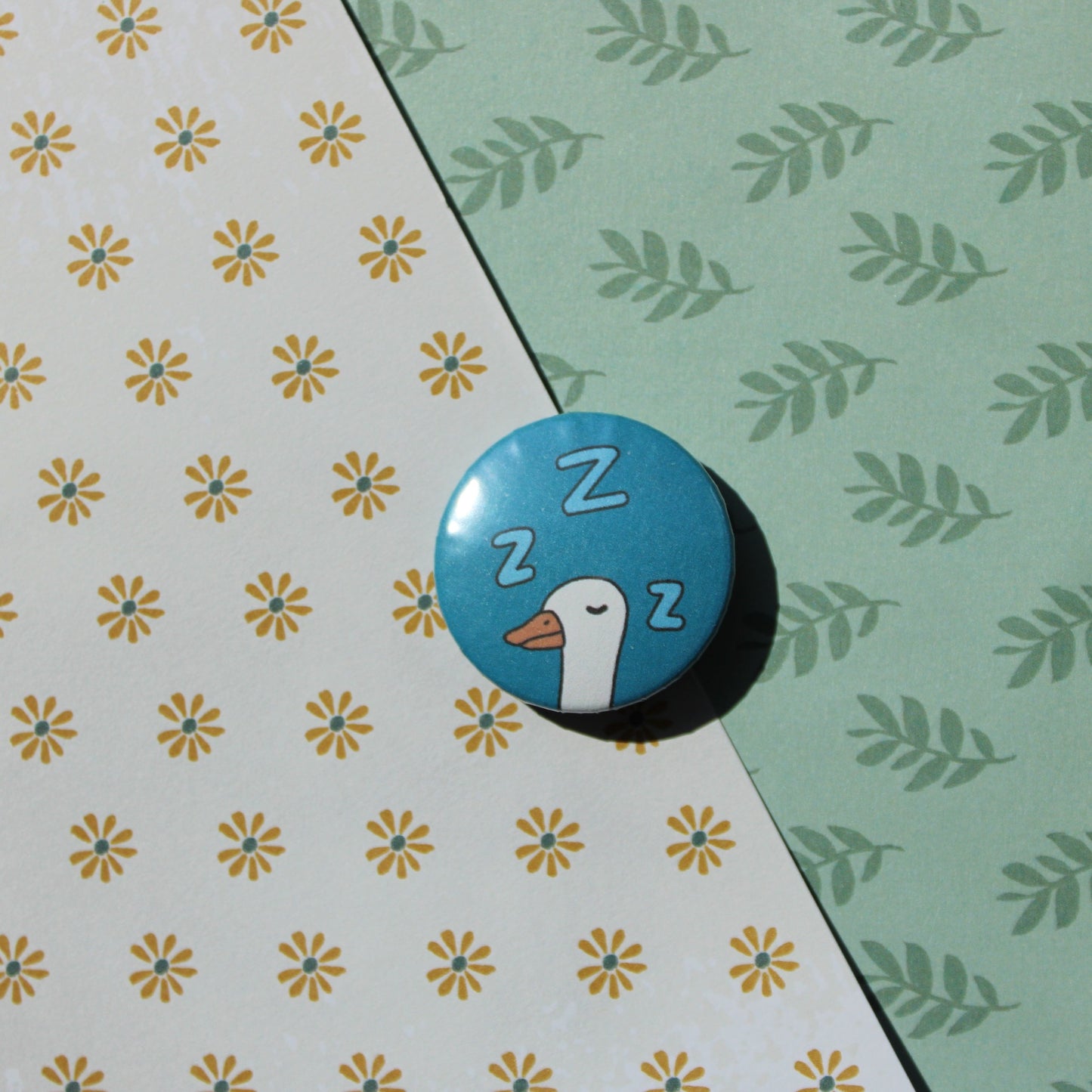 A floral and leaf background with one blue circle button with a white goose head sleeping and three Z's in light blue around it's head. 