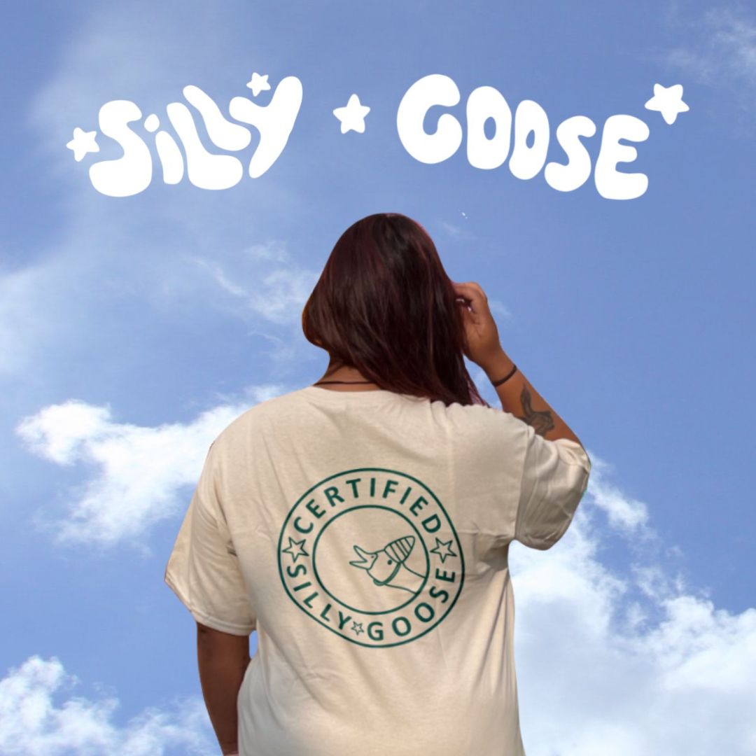 Blue sky background with clouds and "Silly Goose" written on top in white with stars. The back of a woman is seen wearing a beige top with "Certified Silly Goose" and a goose with a party hat popping out in a dark blue outline.