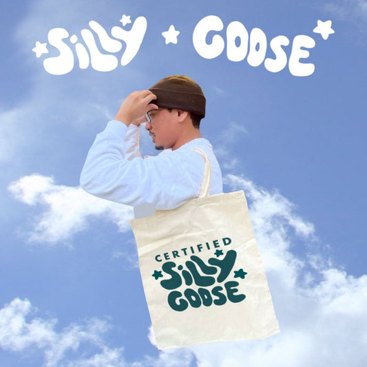 A blue sky with clouds is the background. On the top is written "Silly Goose" with stars. Below is a man holding his beanie floating in the sky with a "Certified Silly Goose" tote bag on his arm. 